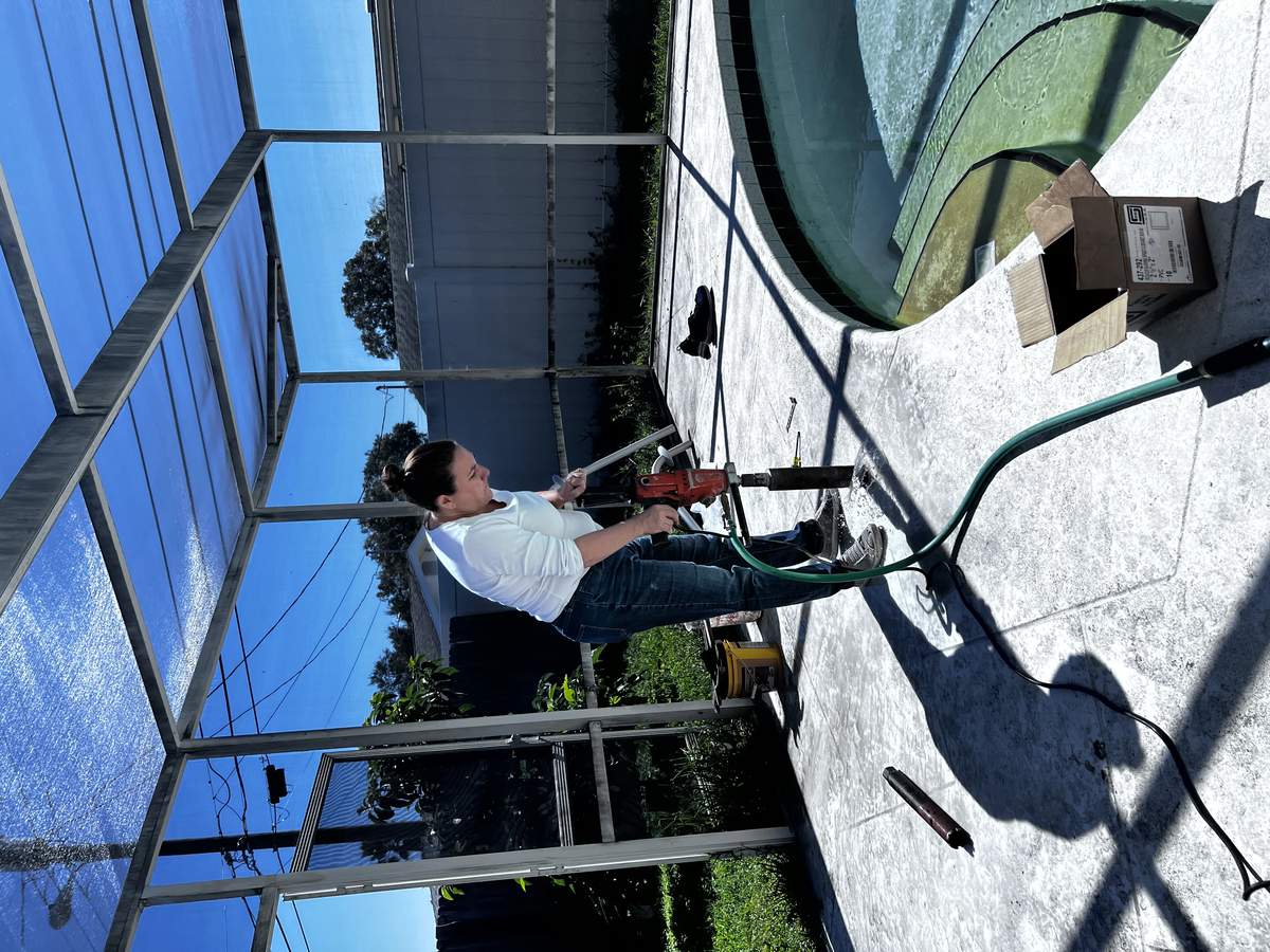 Ada Neikens operating a concrete core drilling machine on a pool deck under a screen